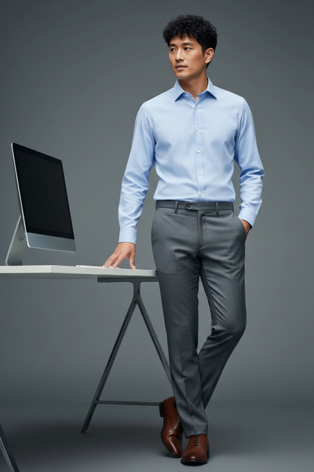 Man in light blue shirt and gray pants standing next to a desk with a computer on a gray background