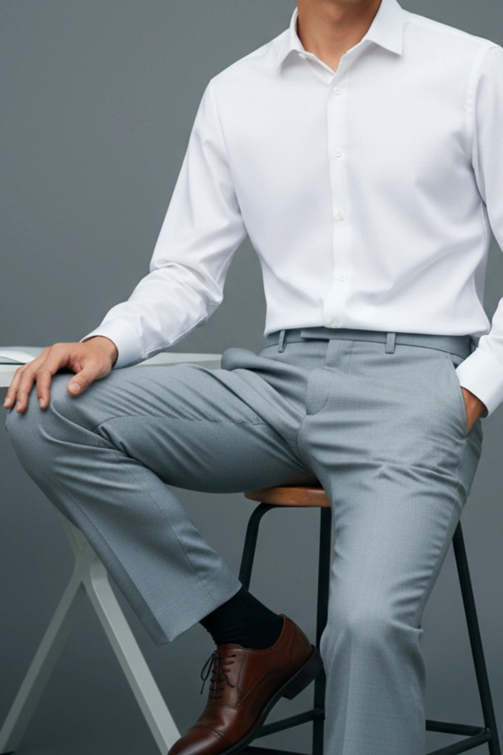 Person wearing a white dress shirt and light gray pants sitting on a stool against a gray background