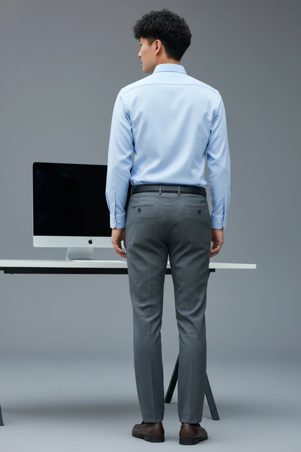 Man in light blue shirt and gray pants standing in front of a computer setup on a gray background