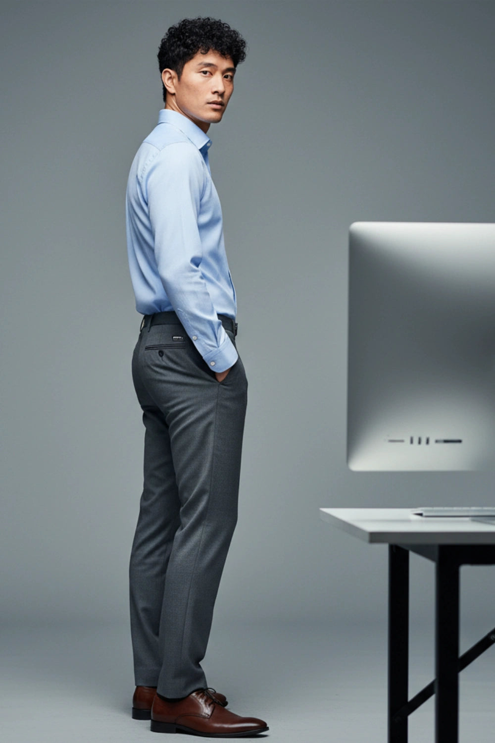 Man in a light blue shirt and gray pants standing next to a computer monitor on a gray background