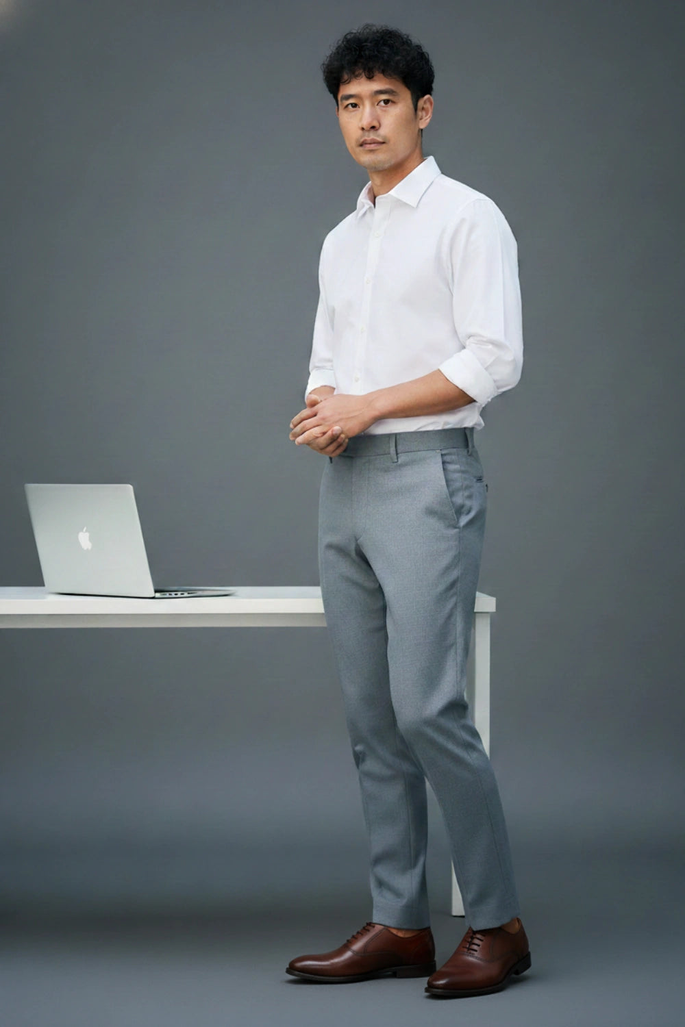 Man in a white shirt and gray pants standing next to a desk with a laptop on a gray background