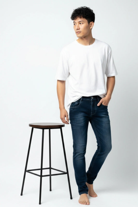 Man wearing a white t-shirt and dark slim fit jeans standing next to a black stool on a white background