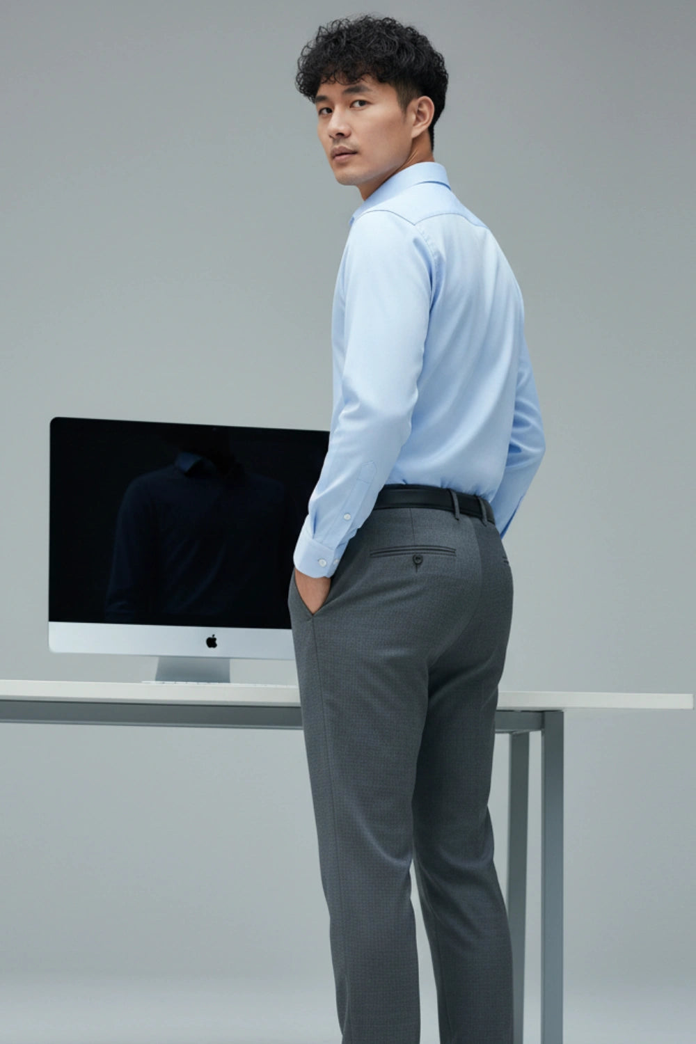 Man in a light blue shirt and gray pants standing in front of a computer monitor.